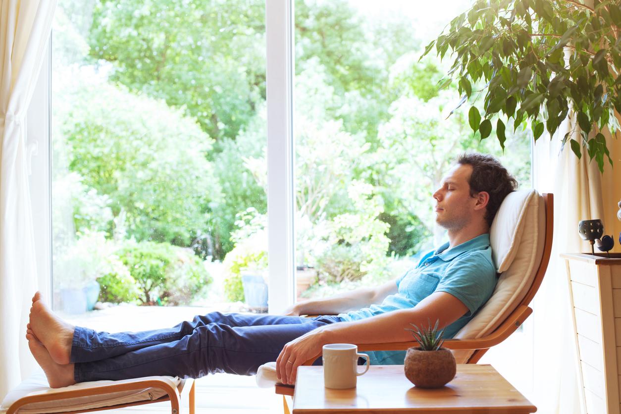 A barefooted man relaxing in his chair in front of a bay window.