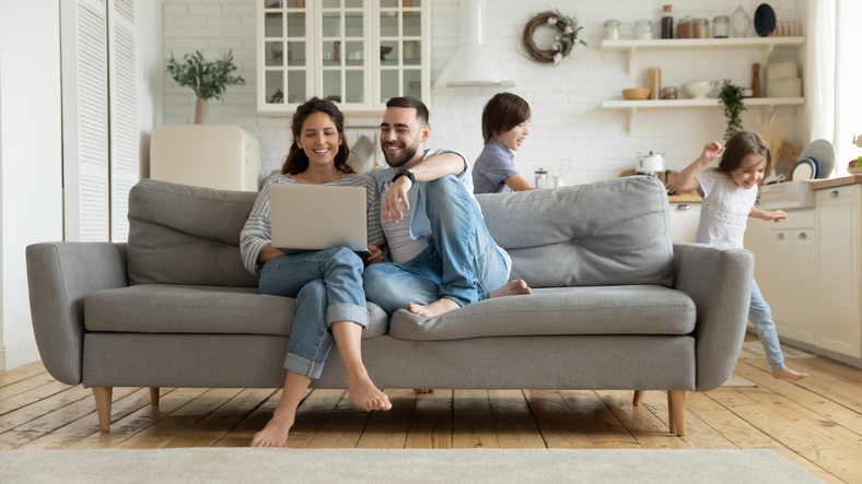 Image of a barefooted couple sitting on their couch looking at something on their laptop, which the wife is holding on her lap, while their children are playing in the background behind the couch.