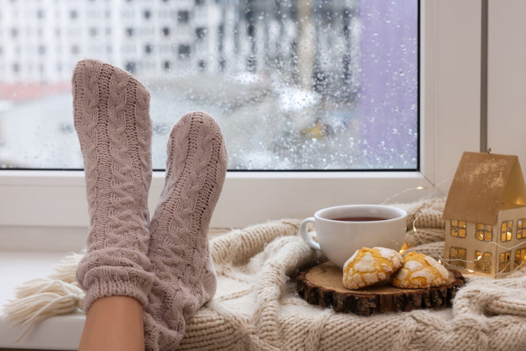 Warm, cozy winter interior scene. A woman's feet are propped on a ledge under a window. She has heavy wool socks on and next to her feet are a cup of coffee and two scones. Raindrops can be seen on the window in the background.
